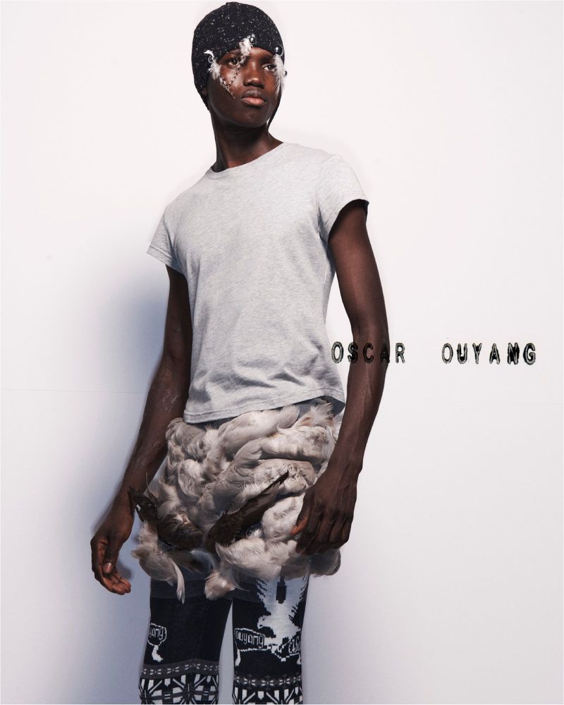 A dark-skinned man in a light gray t-shirt, beanie, and patterned pants poses in a studio with feather accents in a fashion shot.