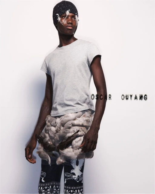 A dark-skinned man in a light gray t-shirt, beanie, and patterned pants poses in a studio with feather accents in a fashion shot.