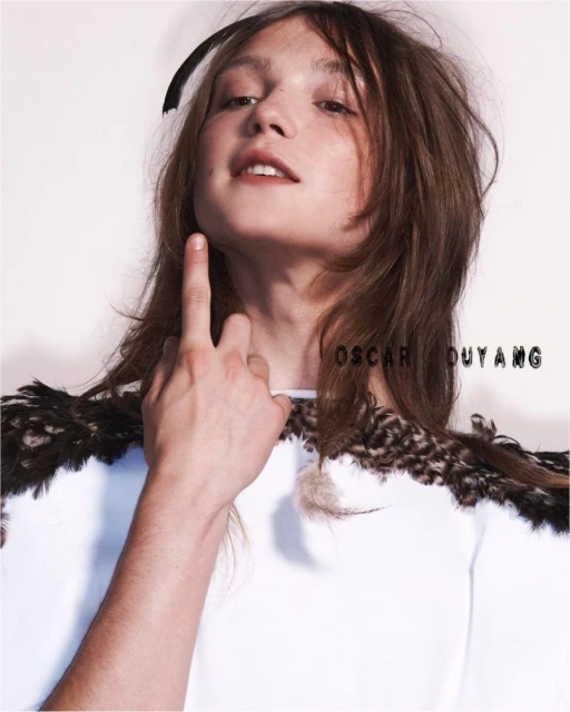 Adult male poses indoors with feather-adorned shirt against a neutral backdrop in a fashion photography setting, with hand raised.