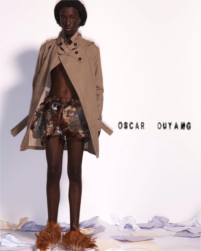 Model wearing an open beige trench coat, textured feathered shorts, and furry brown shoes against a white studio background.
