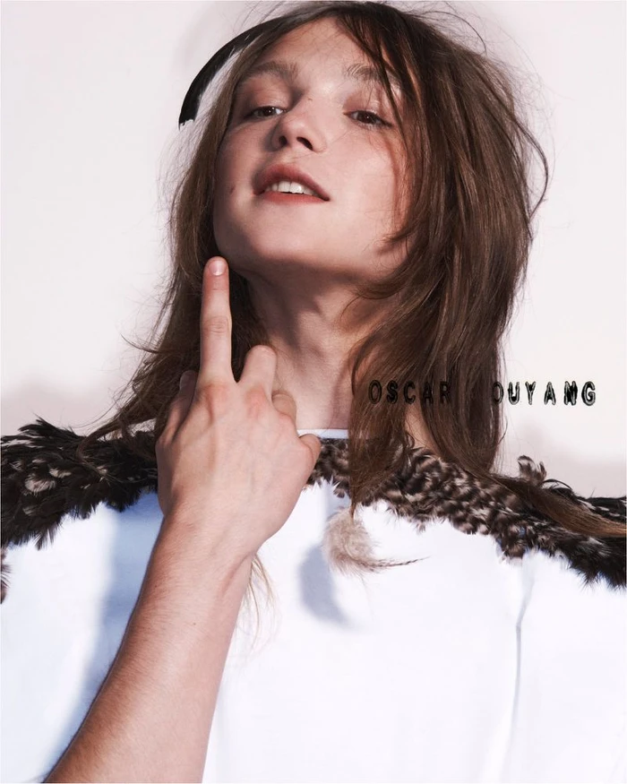 Portrait of a model with long brown hair wearing a white top with feather trim against a plain white background