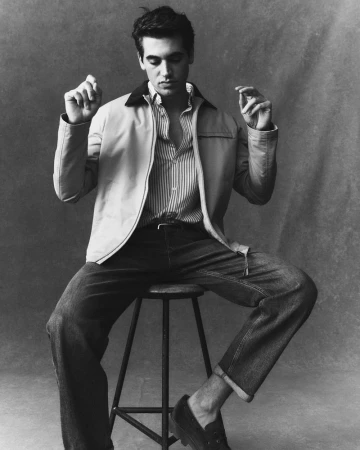 Model Marco Bellotti sitting on a high stool wearing a zip-up jacket, striped shirt, and cuffed jeans against a textured backdrop.