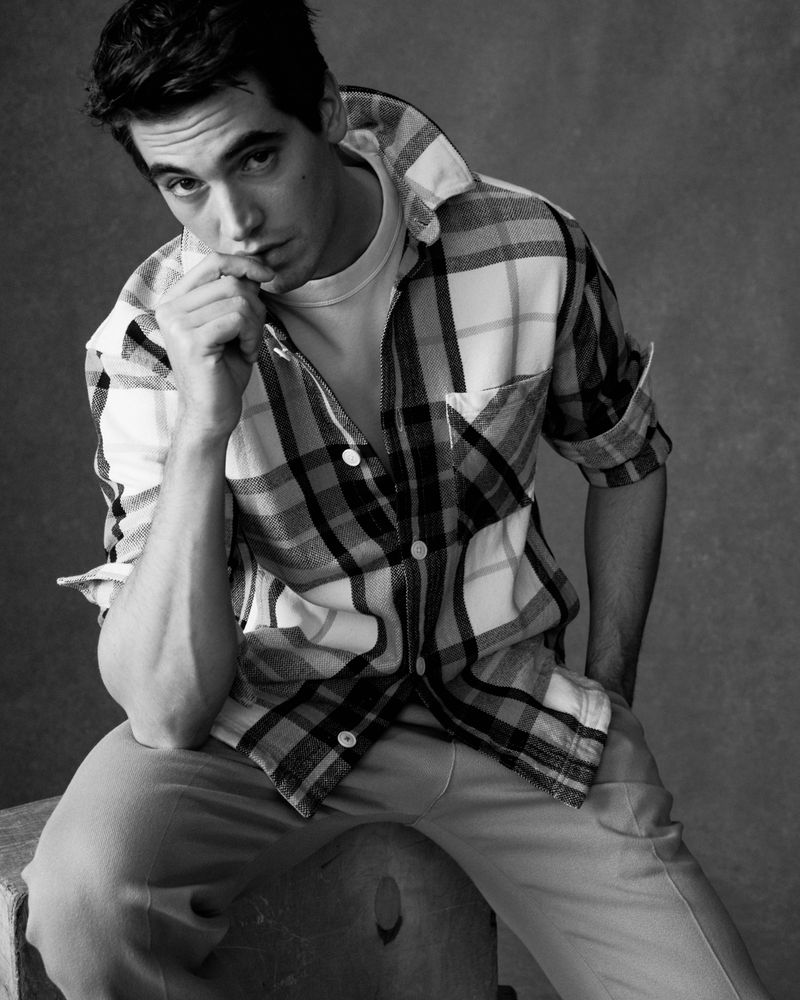 Monochrome portrait of a young adult man in a plaid shirt, seated and pensive, against a neutral background. Focused black-and-white.