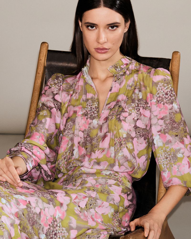 Model Alice Ferazzoli seated in a wooden chair wearing a pink and olive green floral printed long sleeve dress.