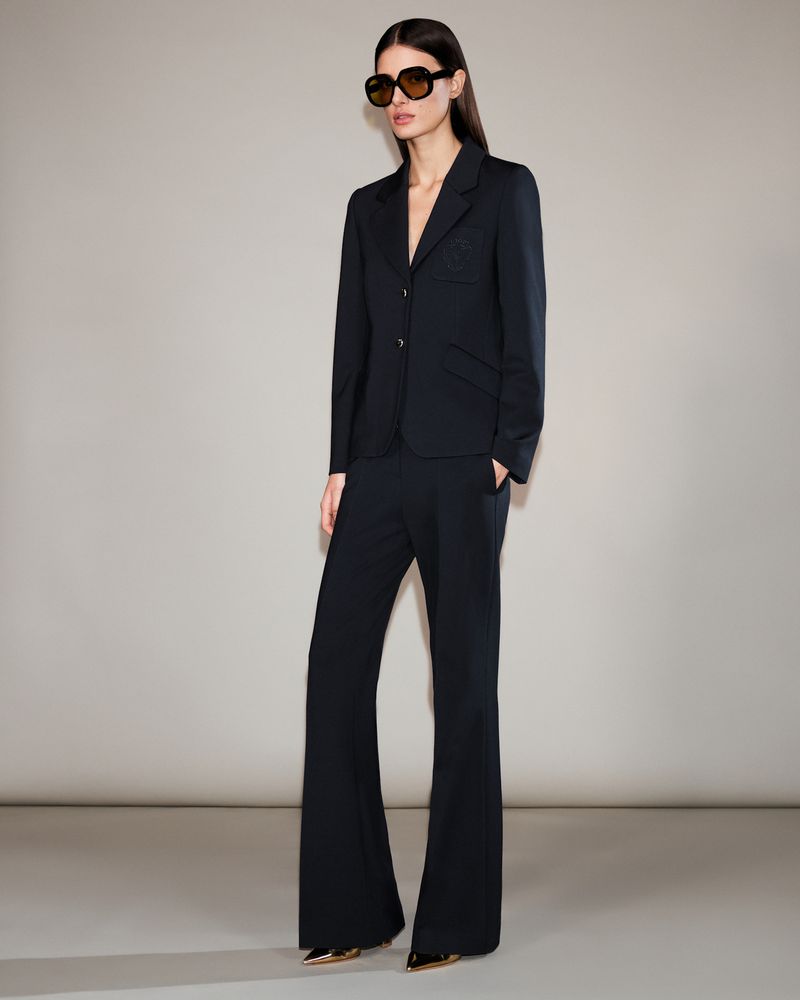 Model wearing dark sunglasses, a black blazer, and matching trousers against a neutral grey studio backdrop.