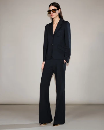 Model wearing dark sunglasses, a black blazer, and matching trousers against a neutral grey studio backdrop.