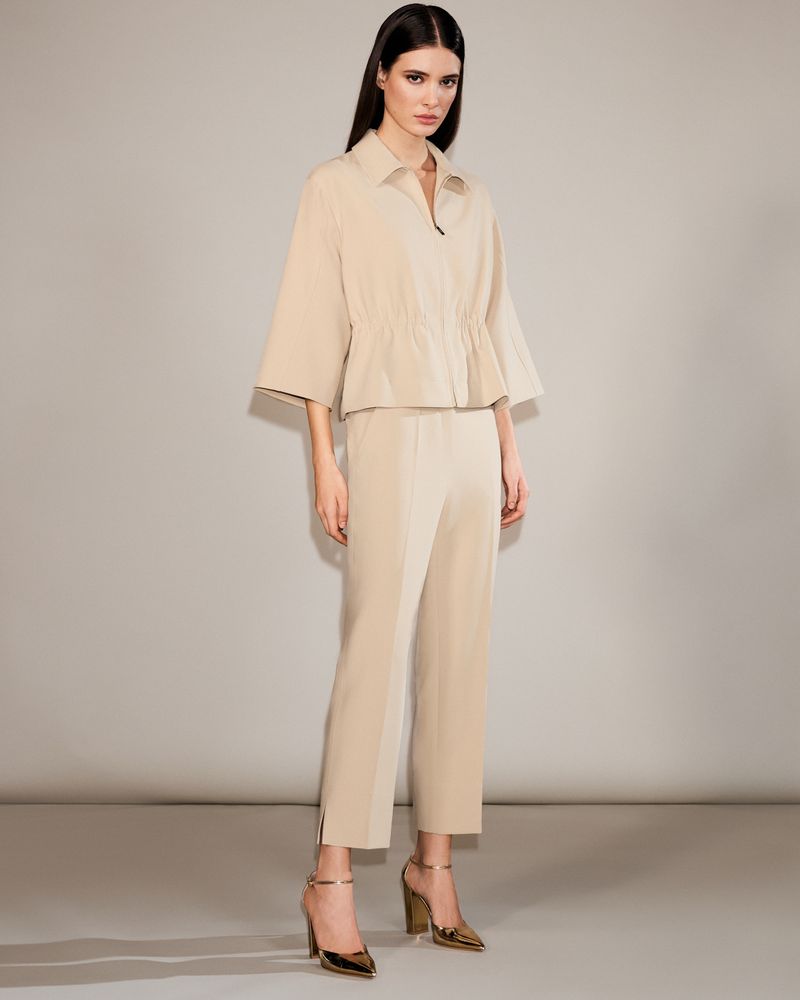 Full-length studio shot of a woman in her twenties wearing a beige jacket, pants, and gold high heels against a light gray backdrop.