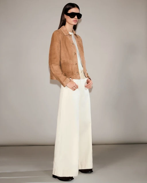 Fashion model poses in a tan jacket and white palazzo pants in a studio. She is wearing black sunglasses and looking at the camera.