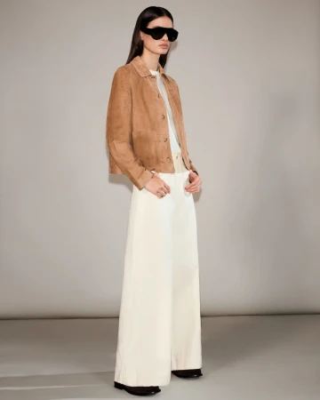 Fashion model poses in a tan jacket and white palazzo pants in a studio. She is wearing black sunglasses and looking at the camera.