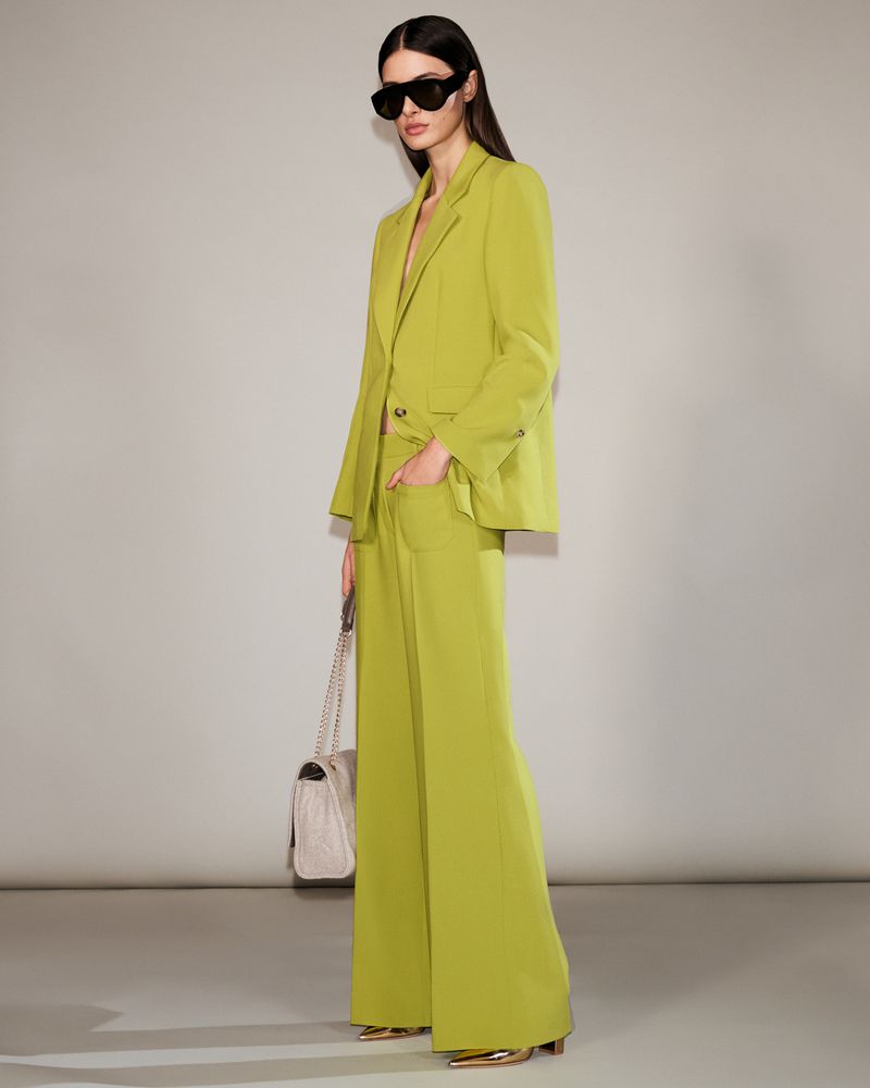 A woman poses in a lime-green suit with a matching handbag, wearing sunglasses, against a neutral backdrop in a fashion studio.