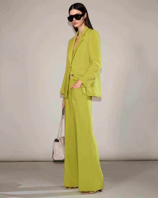 A woman poses in a lime-green suit with a matching handbag, wearing sunglasses, against a neutral backdrop in a fashion studio.
