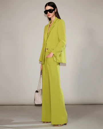 A woman poses in a lime-green suit with a matching handbag, wearing sunglasses, against a neutral backdrop in a fashion studio.