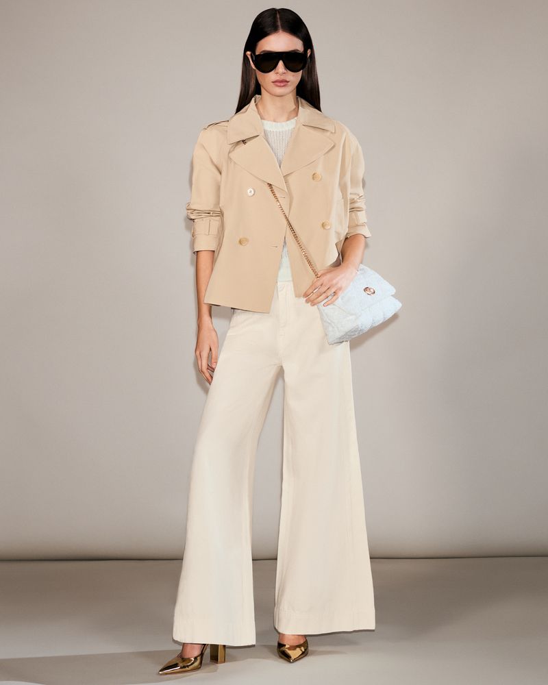 Full-length shot of a woman in a beige cropped trench coat, white wide-leg pants, sunglasses, gold heels, and a cloud-like shoulder bag.