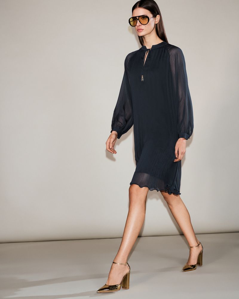 A woman walks in a navy dress and gold heels with a studio background. She is also wearing sunglasses, and the shot is medium.