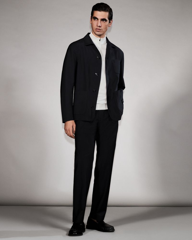 Model Marco Bellotti wearing a black jacket, white zip-neck top, and black trousers against a neutral grey studio background.