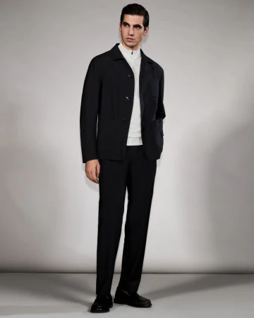 Model Marco Bellotti wearing a black jacket, white zip-neck top, and black trousers against a neutral grey studio background.