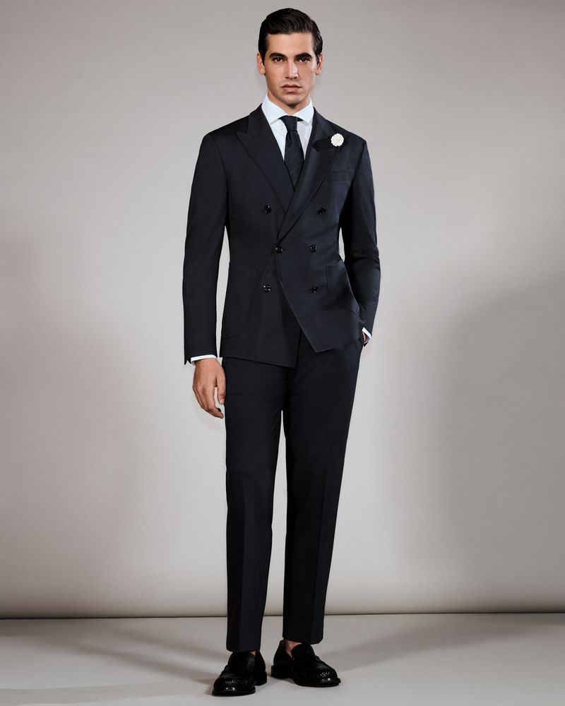 Elegant adult male stands in a black double-breasted suit, white dress shirt, and tie against a neutral studio backdrop.