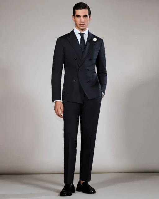 Elegant adult male stands in a black double-breasted suit, white dress shirt, and tie against a neutral studio backdrop.