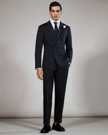 Elegant adult male stands in a black double-breasted suit, white dress shirt, and tie against a neutral studio backdrop.