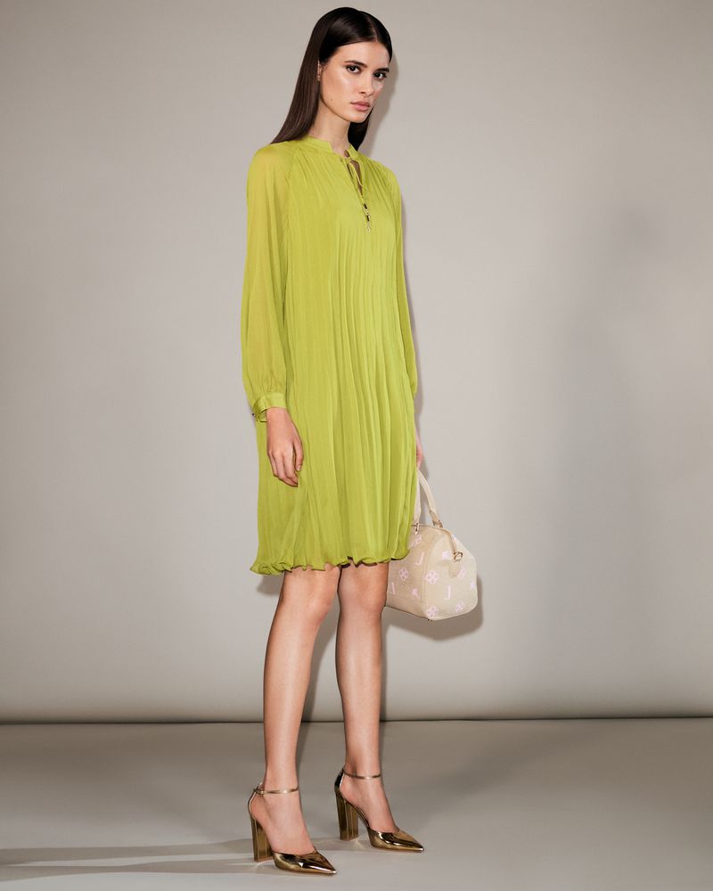 Model Alice Ferazzoli wearing a lime green pleated long-sleeve dress and metallic gold heels holding a handbag against a studio backdrop.
