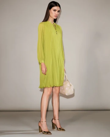 Model Alice Ferazzoli wearing a lime green pleated long-sleeve dress and metallic gold heels holding a handbag against a studio backdrop.