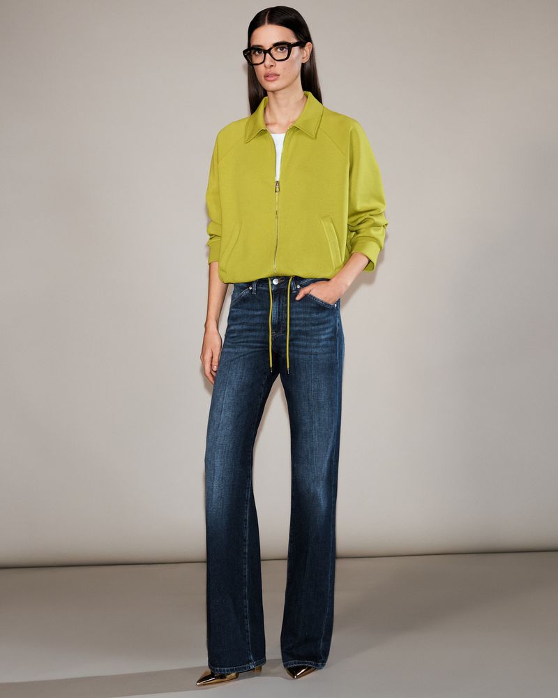 Full-length portrait of an adult woman with glasses, wearing a chartreuse jacket and blue jeans, posing in front of a solid grey backdrop.