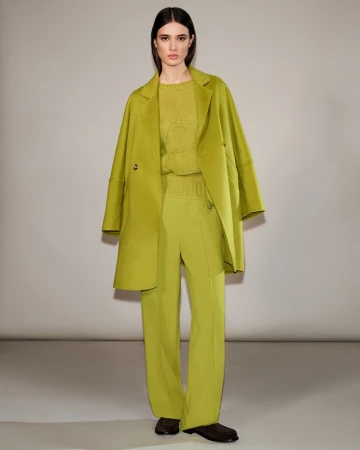 A woman wears a chartreuse wool coat over a matching knit top and flared pants, complemented by dark brown shoes, posing indoors.