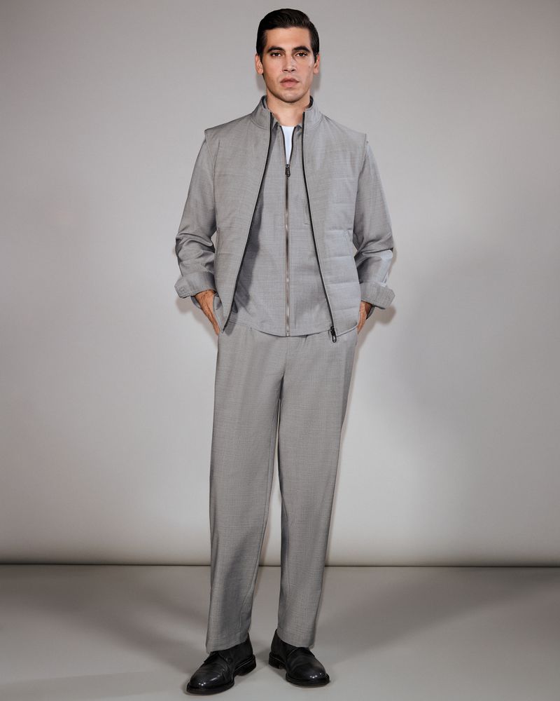 Full-length studio shot of a young adult male model wearing a gray casual suit. He poses with his hands in his pockets against a plain backdrop.