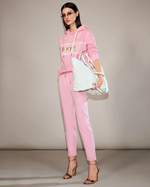 A woman models a pink patterned hooded sweatshirt with matching pants, gold heels, and sunglasses in a studio setting, holding a white tote bag.