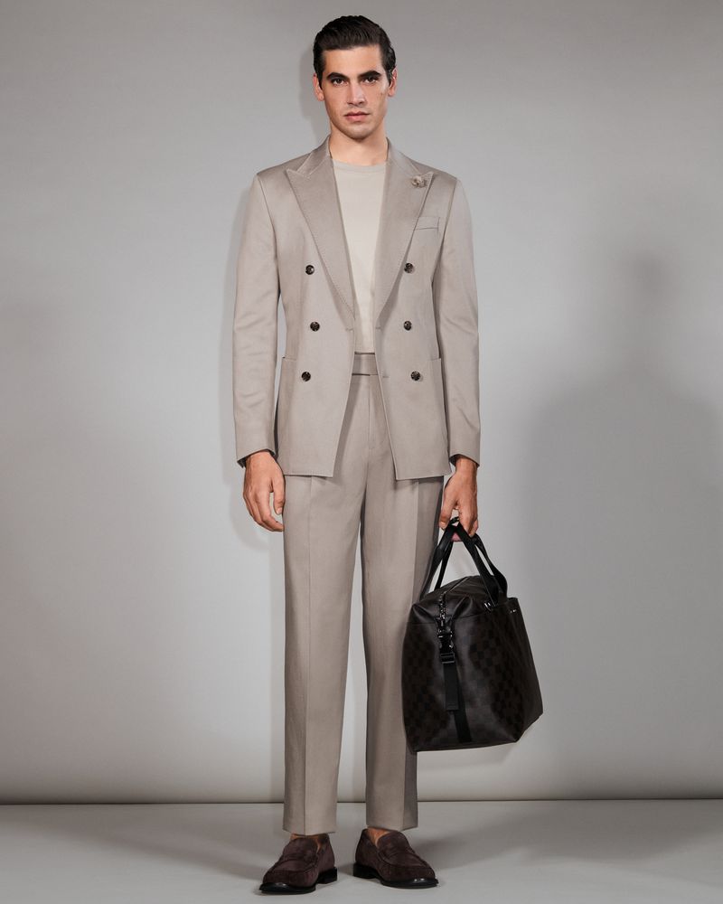 Man in a suit stands in a full shot, holding a duffel bag. He wears brown loafers and poses in front of a grey backdrop.