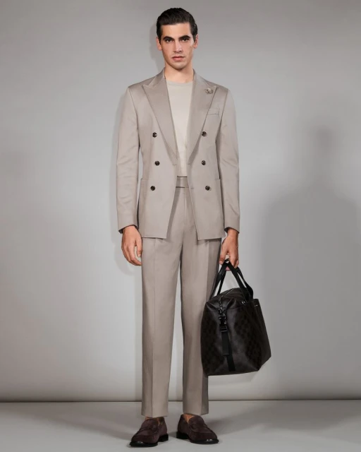 Man in a suit stands in a full shot, holding a duffel bag. He wears brown loafers and poses in front of a grey backdrop.