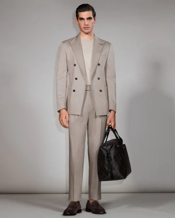 Man in a suit stands in a full shot, holding a duffel bag. He wears brown loafers and poses in front of a grey backdrop.