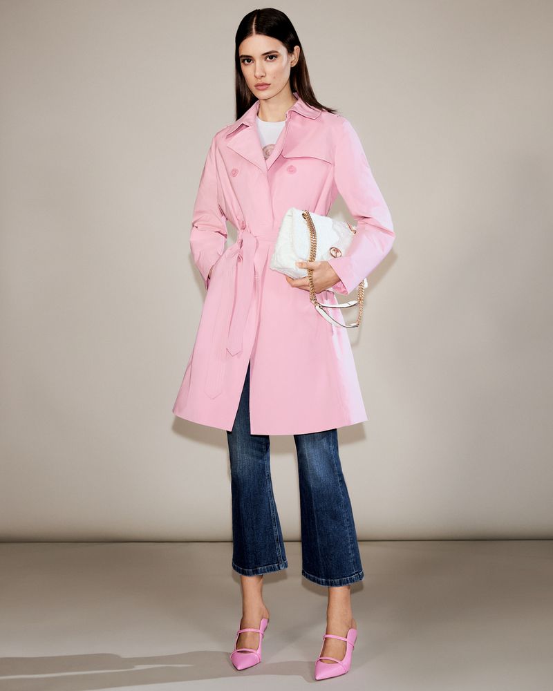 Elegant woman in pink trench coat, cropped blue jeans, and pink heels, holding a white purse, posing in a neutral studio setting.