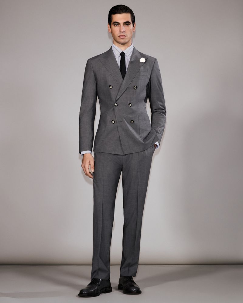 A man stands in a photo studio, wearing a grey double-breasted suit with a black tie, presenting a formal and fashionable appearance.