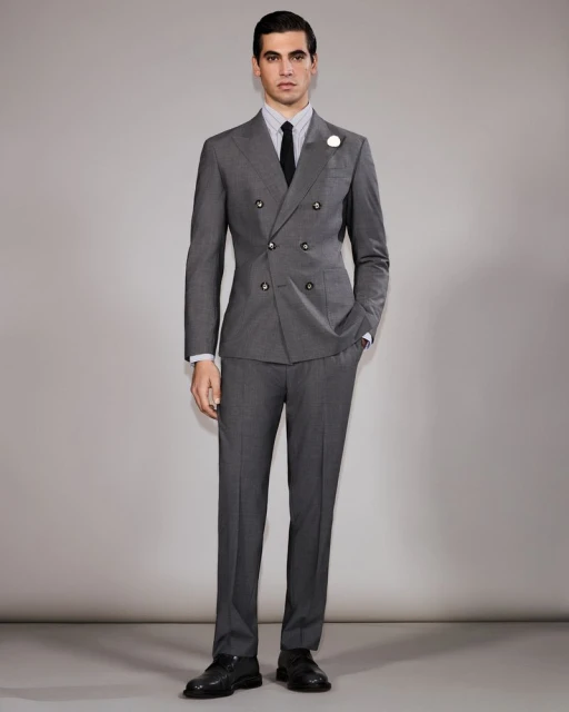A man stands in a photo studio, wearing a grey double-breasted suit with a black tie, presenting a formal and fashionable appearance.