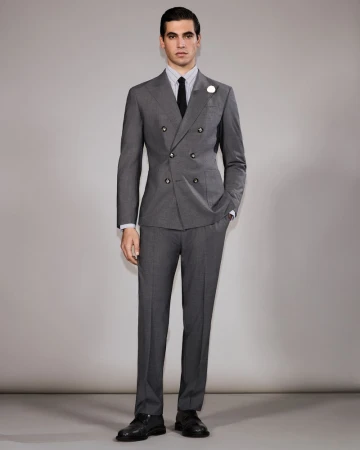 A man stands in a photo studio, wearing a grey double-breasted suit with a black tie, presenting a formal and fashionable appearance.