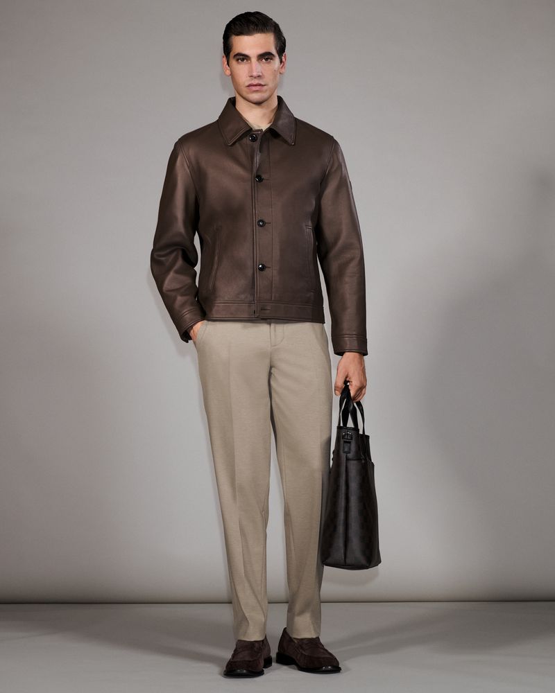 Man in brown leather jacket, beige pants, and brown shoes holding a dark brown tote bag stands in a neutral studio setting.