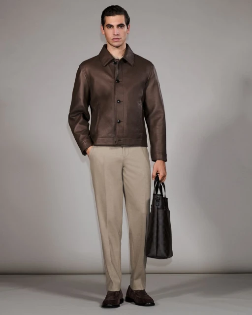Man in brown leather jacket, beige pants, and brown shoes holding a dark brown tote bag stands in a neutral studio setting.