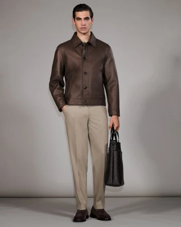 Man in brown leather jacket, beige pants, and brown shoes holding a dark brown tote bag stands in a neutral studio setting.