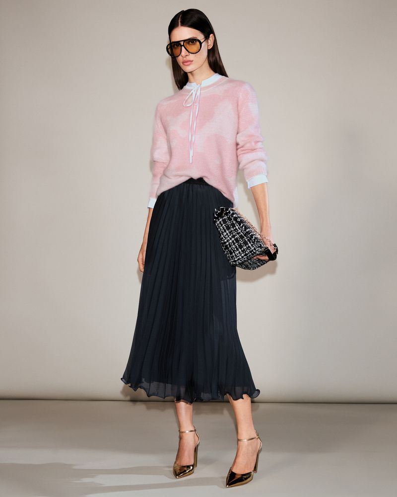 Full-length shot of a woman in a pink sweater, black pleated skirt, sunglasses, and gold heels, holding a small tweed handbag, indoors.