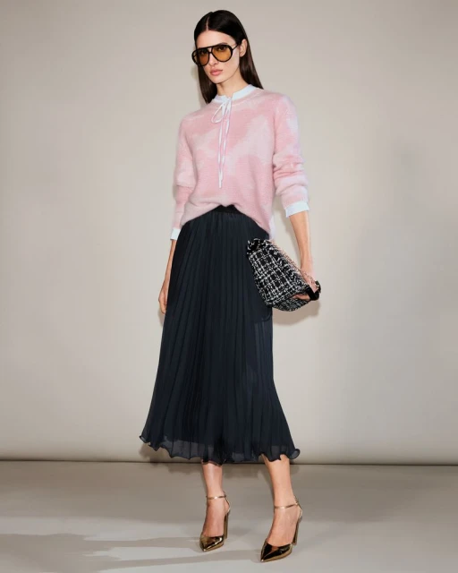 Full-length shot of a woman in a pink sweater, black pleated skirt, sunglasses, and gold heels, holding a small tweed handbag, indoors.