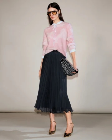 Full-length shot of a woman in a pink sweater, black pleated skirt, sunglasses, and gold heels, holding a small tweed handbag, indoors.