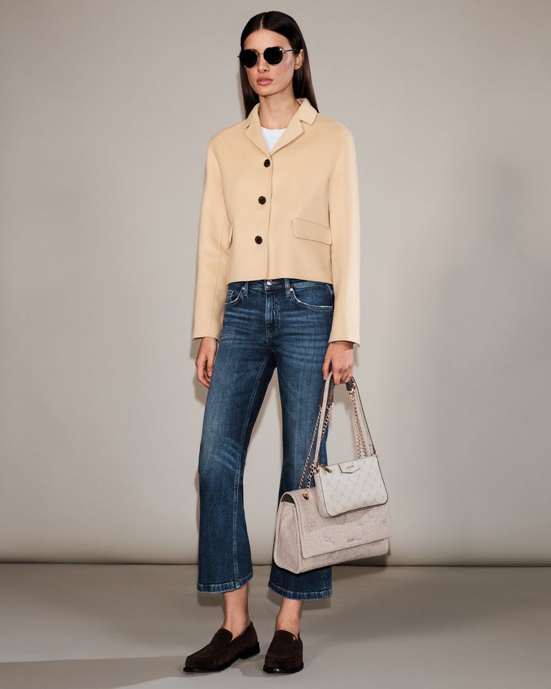 A woman models a cropped beige blazer, dark wash jeans, dark loafers, and a light quilted shoulder bag in a studio setting.