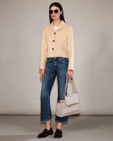 A woman models a cropped beige blazer, dark wash jeans, dark loafers, and a light quilted shoulder bag in a studio setting.