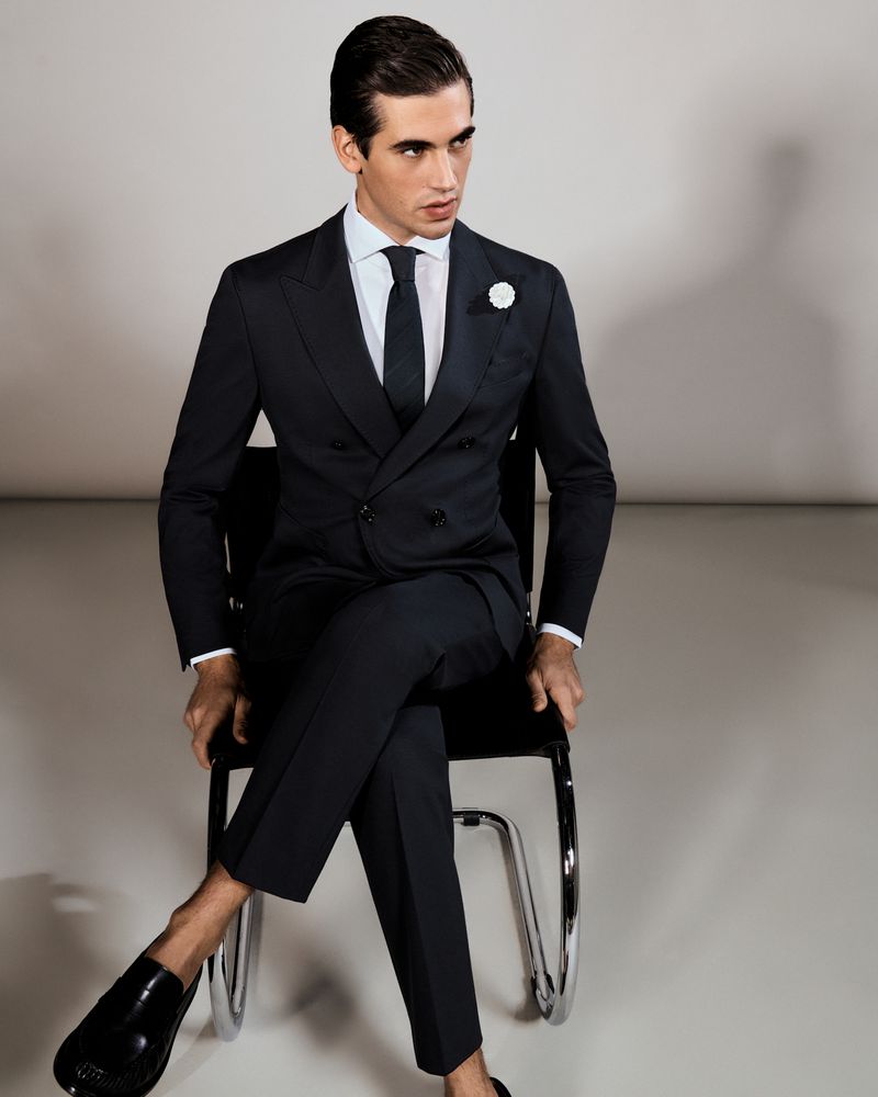 A handsome adult male in a formal dark suit sits on a chair in a bright photo studio, modeling the suit and wearing shiny black shoes.