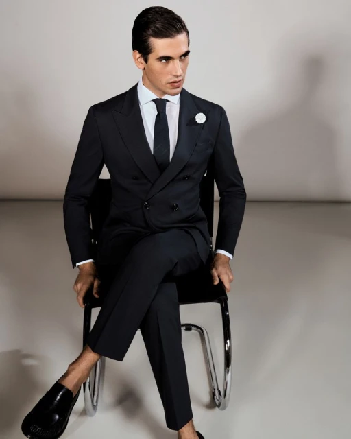 A handsome adult male in a formal dark suit sits on a chair in a bright photo studio, modeling the suit and wearing shiny black shoes.