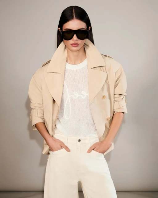 A brunette woman wearing a light-tan trench coat and white jeans poses against a neutral backdrop with dark sunglasses and hands in pockets.