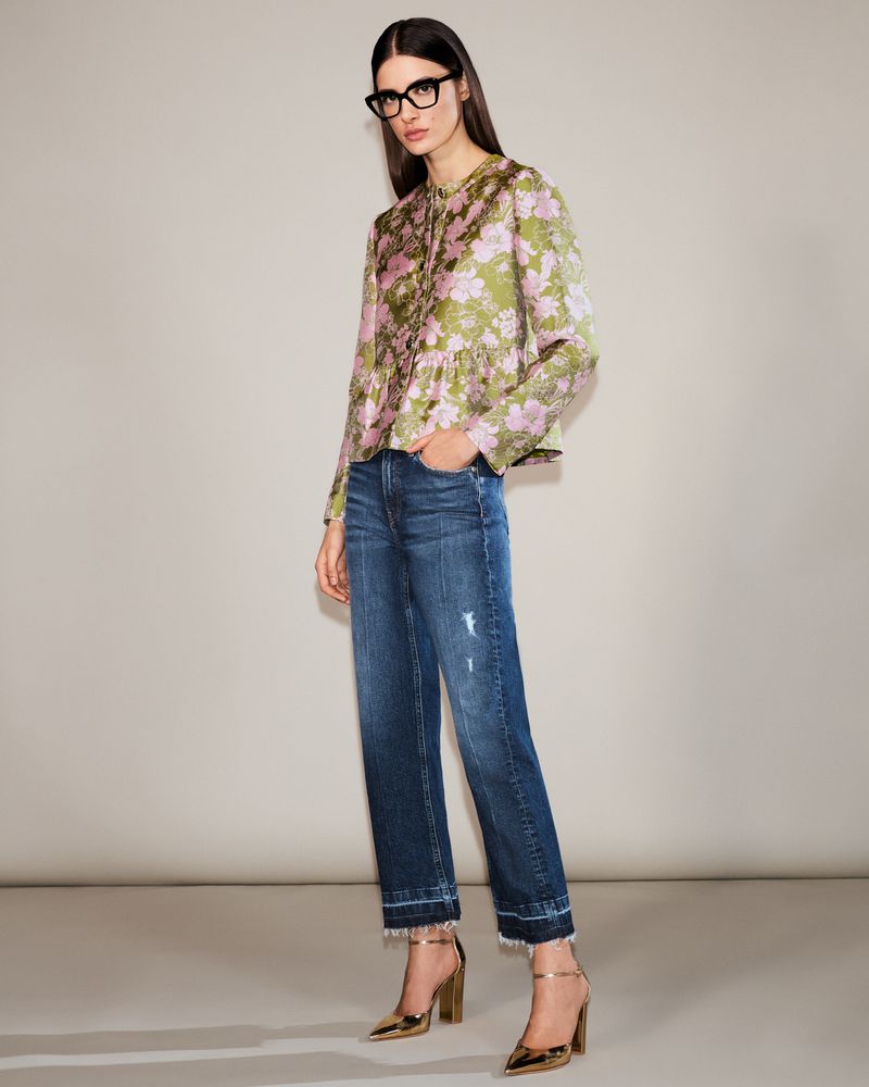 A woman models a floral top, blue jeans, and gold heels in a studio setting, full shot. Fashionable attire for women.
