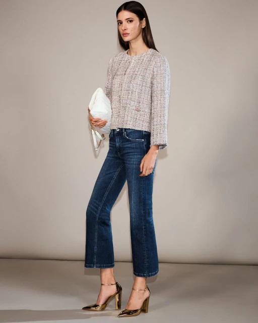 Fashionable woman in tweed jacket, dark wash jeans, and gold high heels, carrying a white clutch bag, posing in a studio setting.
