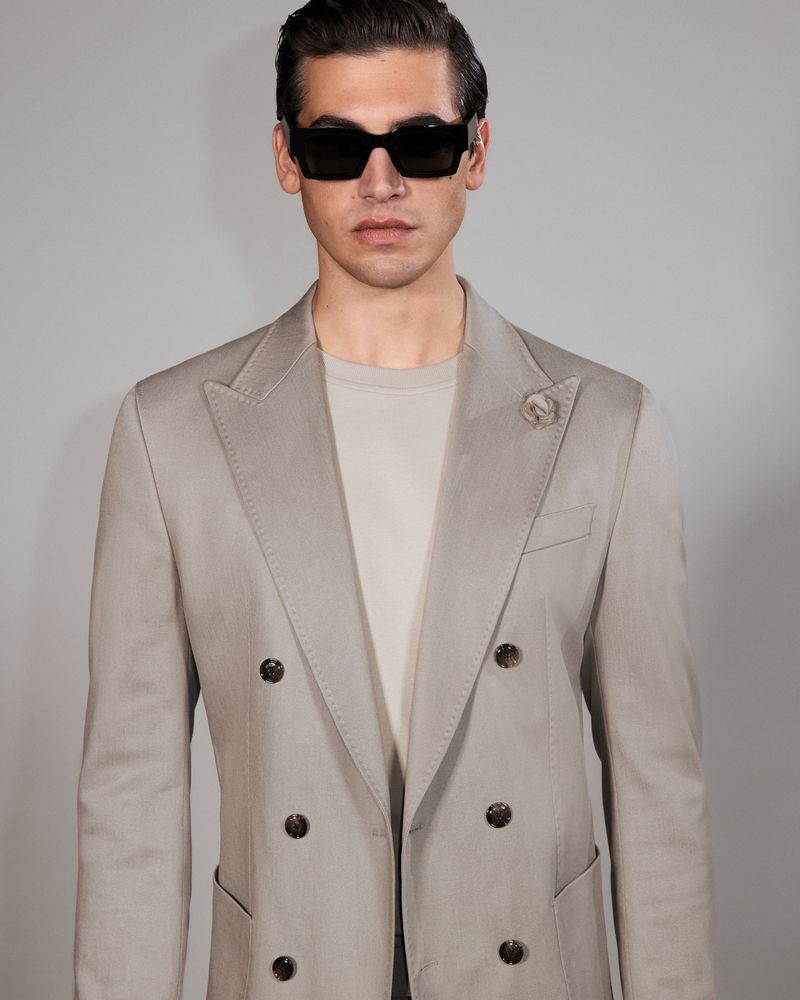 Portrait of a handsome man with dark hair and sunglasses, wearing a double-breasted blazer with a lapel pin, posing against a light backdrop.